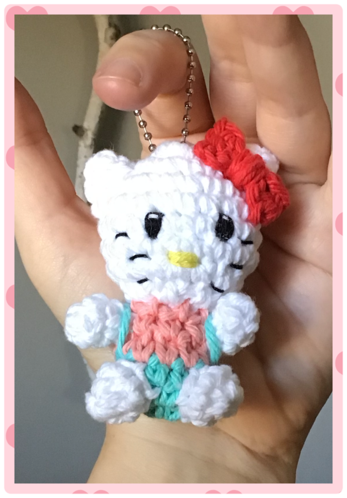 My hand holding up a tiny hello kitty stuffie in a keychain. She is in pink t-shirt and teal overalls with her big red bow at the ear