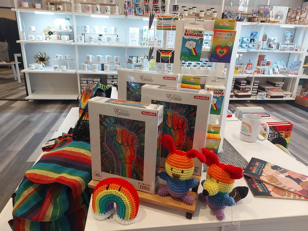 Picture of the inside of the shop. Shelves with swag in the background. The foreground is a table with pride swag from mugs, puzzles and my stuffies!