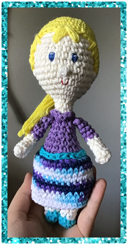 A crochet stuffie doll in a top and skirt. She is blond and blue eyed
