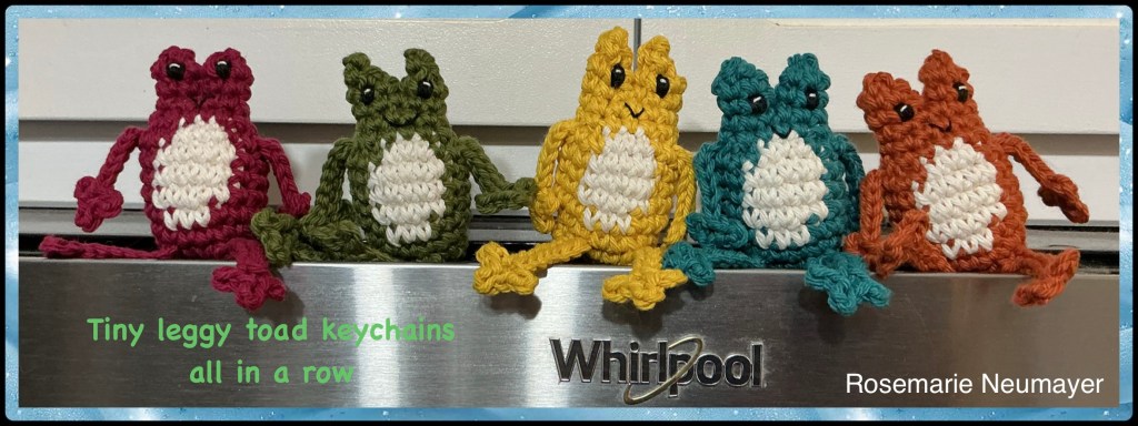 Tiny leggy toad keychains all in a row on top of my microwave. Left to right are burgundy, sage green, gold, teal, and turmeric coloured toads